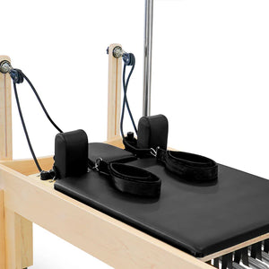 Pilates reformer machine with black cushion and straps on a wooden frame.