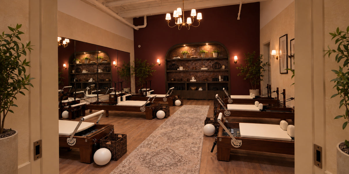 Two rows of solid American walnut Oshun Timber Reformers with ivory upholstery in a boutique Pilates studio with an oxblood wall and a brass chandelier