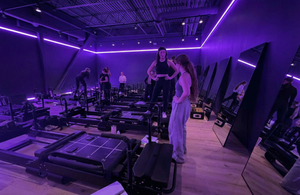 Gym interior with Pilates reformers and people exercising under purple lighting