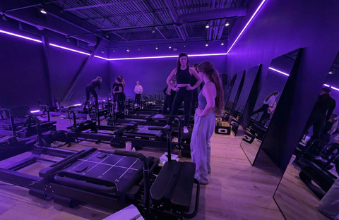 Gym interior with Pilates reformers and people exercising under purple lighting