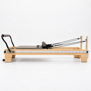 Side view of the Oshun Timber Therapy Reformer in maple, with the full-length footbar rail and rope risers