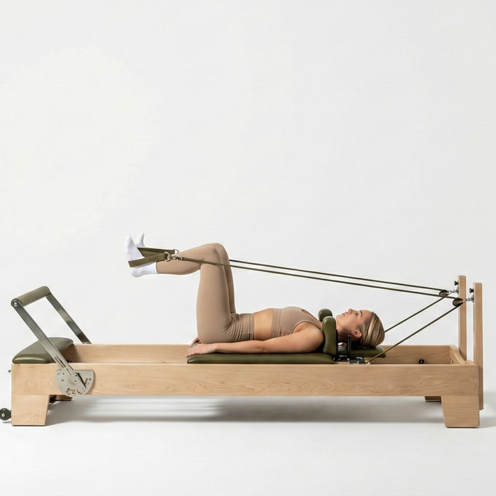 Woman doing a feet-in-straps exercise on an Oshun Timber Reformer in maple with olive green upholstery