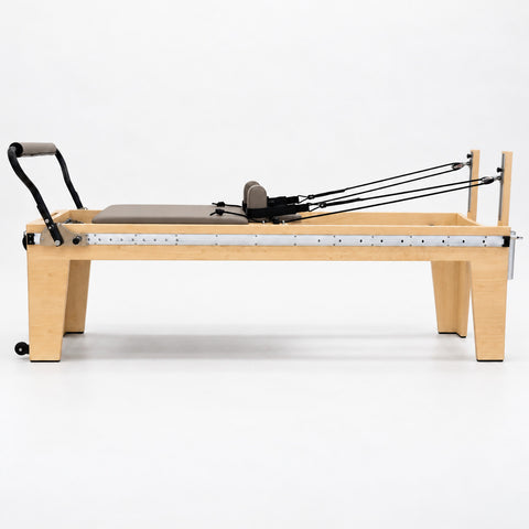 Side view of the Oshun Timber Therapy Reformer at Rehab Height, a 24 in maple frame on tall tapered legs, with the full-length footbar rail